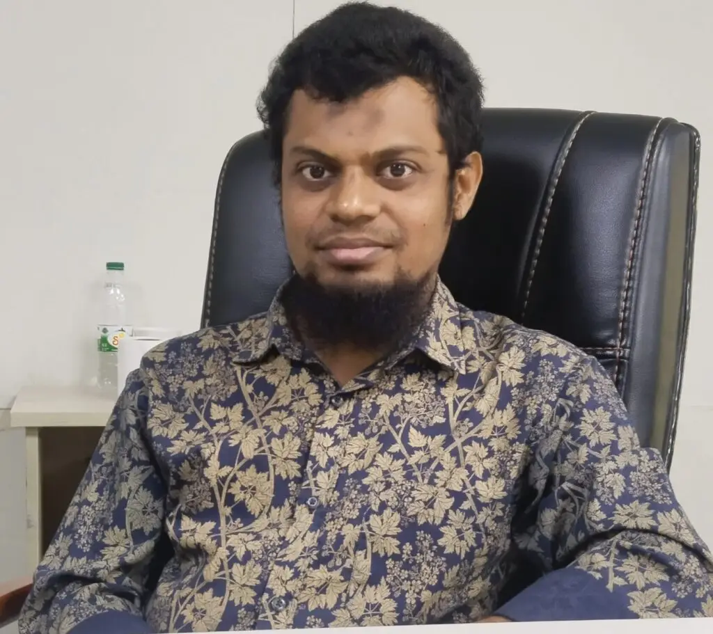 Mizanur Rahman, a young man with short curly black hair and a beard, wearing a navy blue floral shirt, standing against a plain white background with a confident smile.